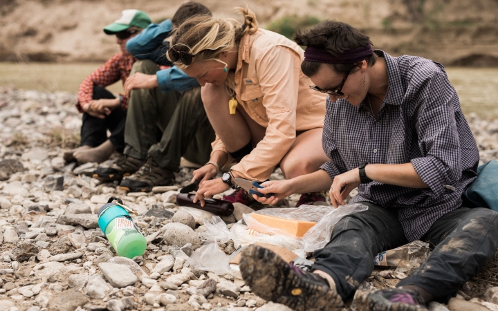four people sit on a rocky shore and prepare food